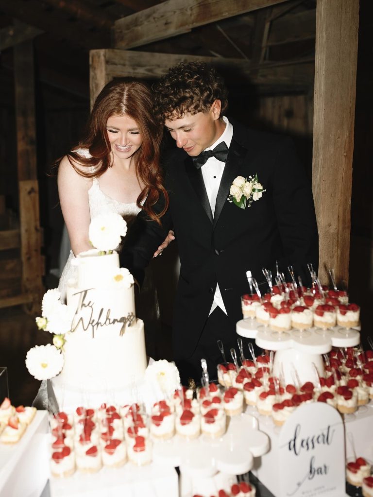 Bride and groom cutting white wedding cake with gold calligraphy topper and fresh flower decorations alongside strawberry dessert bar