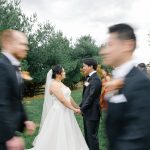 Newlyweds hold hands on lawn as wedding party in motion blurs past them during outdoor fall celebration