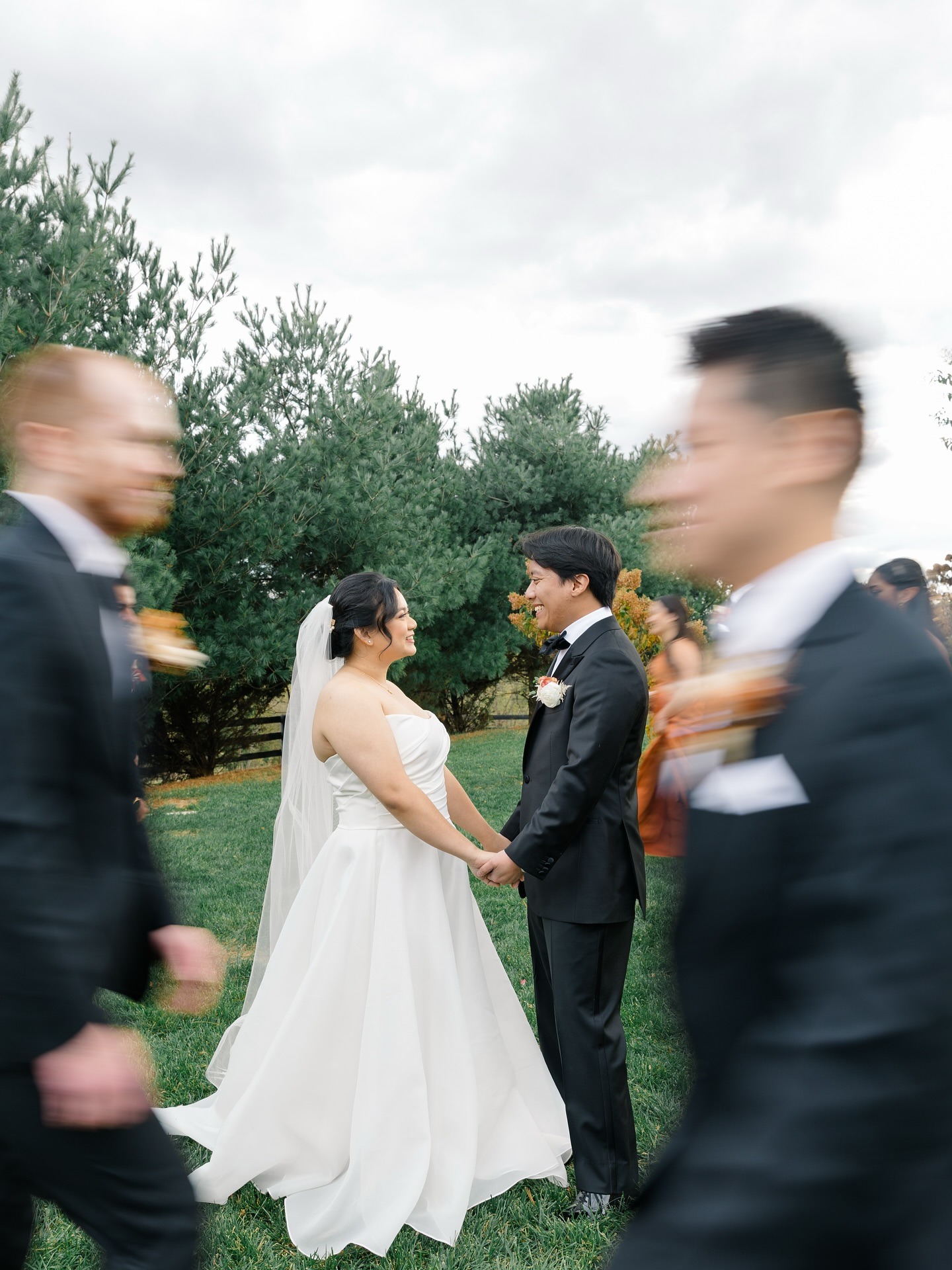 Newlyweds hold hands on lawn as wedding party in motion blurs past them during outdoor fall celebration