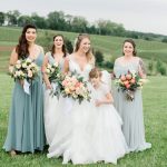 Bride with flower girl and bridesmaids in sage and gray gowns holding pastel bouquets on vineyard hillside