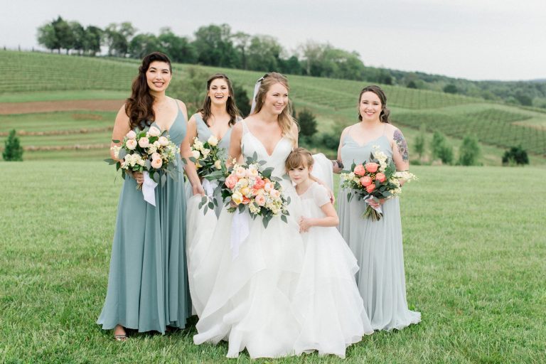 Bride with flower girl and bridesmaids in sage and gray gowns holding pastel bouquets on vineyard hillside