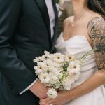 Bride holding white bouquet with shoulder tattoo embraces groom in dark suit