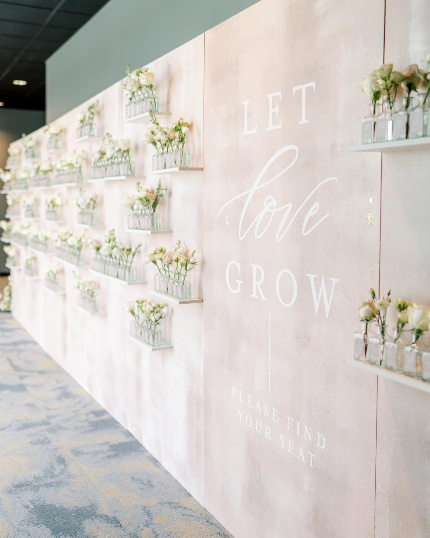 Wedding escort card display wall in soft blush tones with rows of bud vases and romantic calligraphy signage