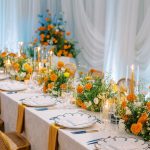 Wedding reception table with cane-back chairs, blue-trimmed china, orange and white floral arrangements, and tall pillar candles