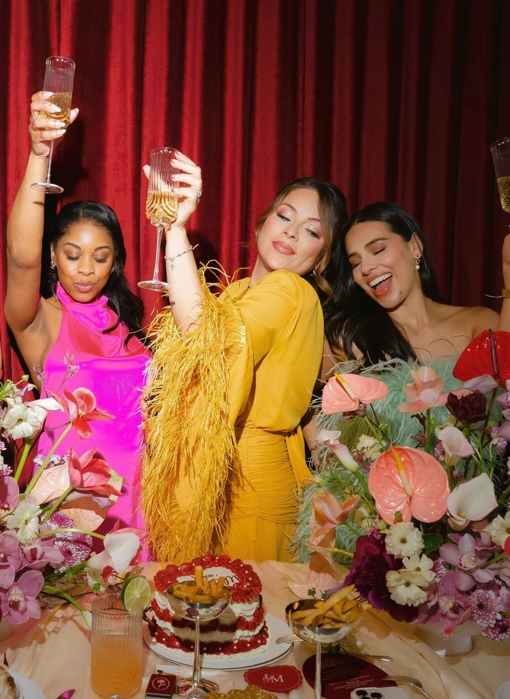 Three women in yellow, pink, and magenta dresses celebrating with arms raised in front of red curtain and colorful flowers