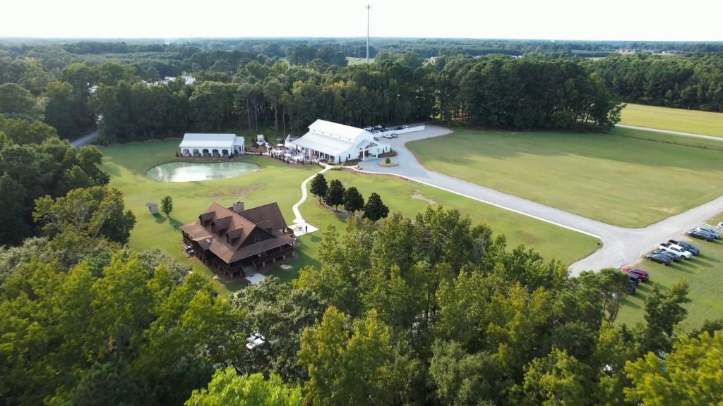 Aerial view of The Hartwood estate featuring timber lodge, white event tent, pond, and surrounding woodlands