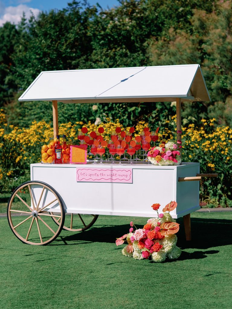 Spritz cart stationed on lawn with colorful drinks and floral decorations surrounded by yellow blooms and greenery