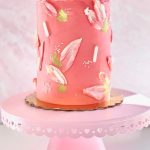 Coral buttercream wedding cake decorated with pink fondant brushstrokes, gold leaf accents, and pearl dots on pink cake stand