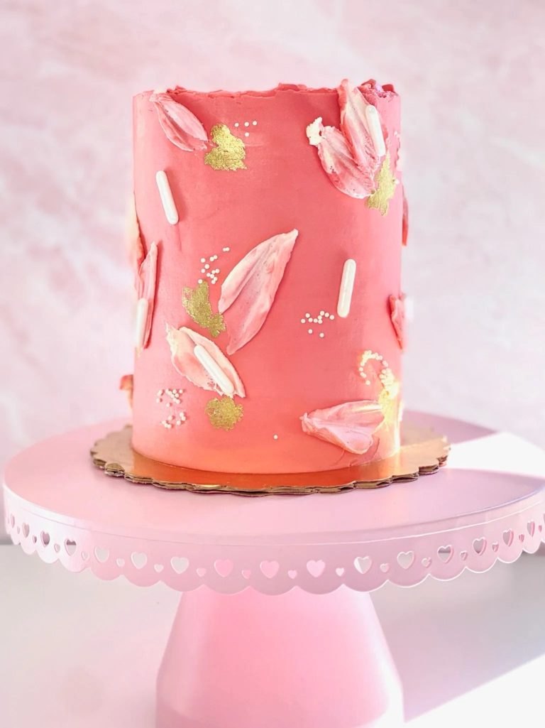 Coral buttercream wedding cake decorated with pink fondant brushstrokes, gold leaf accents, and pearl dots on pink cake stand