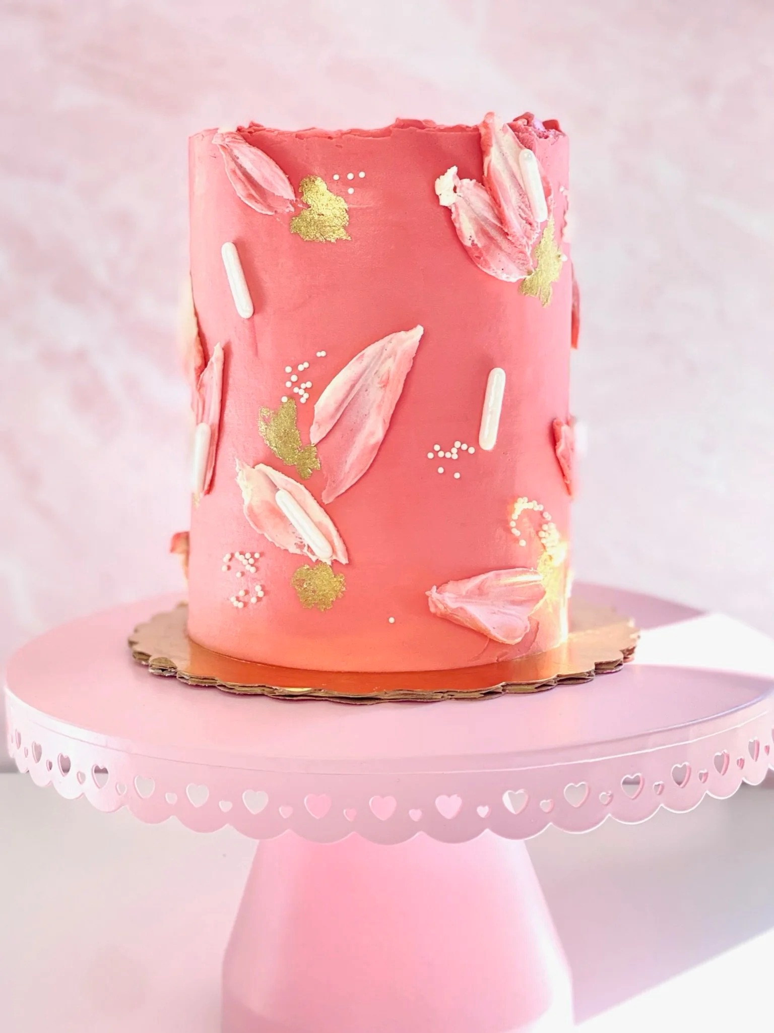 Coral buttercream wedding cake decorated with pink fondant brushstrokes, gold leaf accents, and pearl dots on pink cake stand