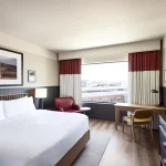 Bright hotel suite with premium bedding, distinctive scarlet valances, comfortable lounge chair, and urban views
