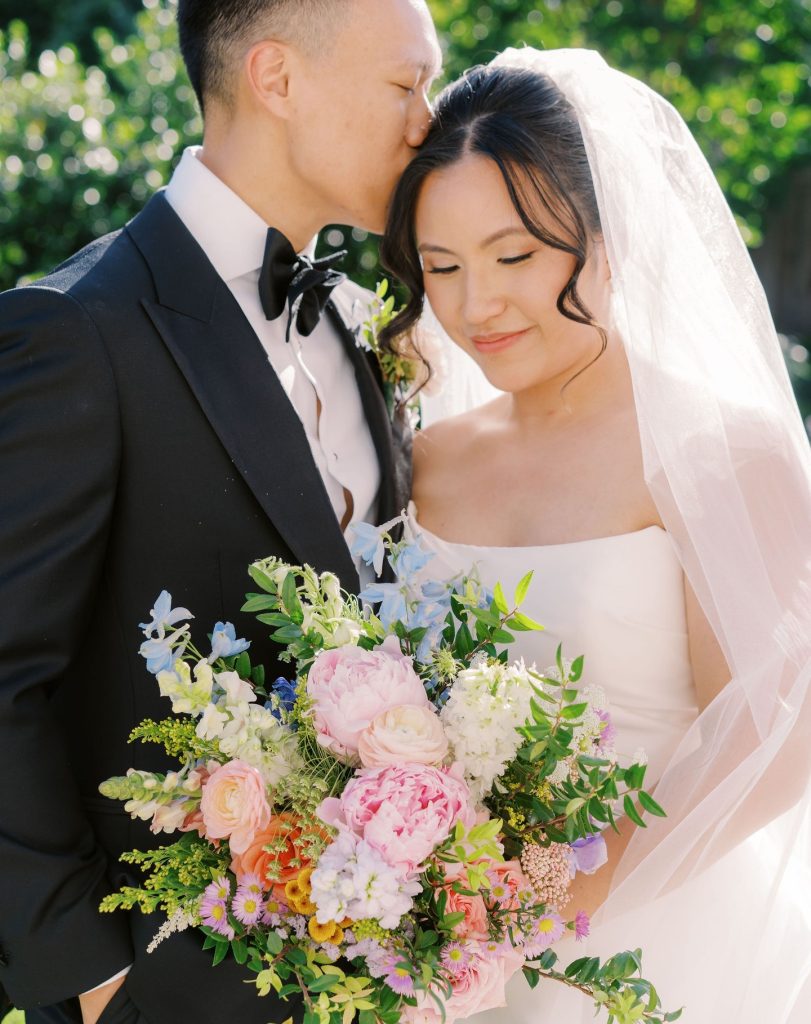 Bride holding lush bouquet of pink peonies, roses, blue delphiniums, and greenery with groom kissing her forehead