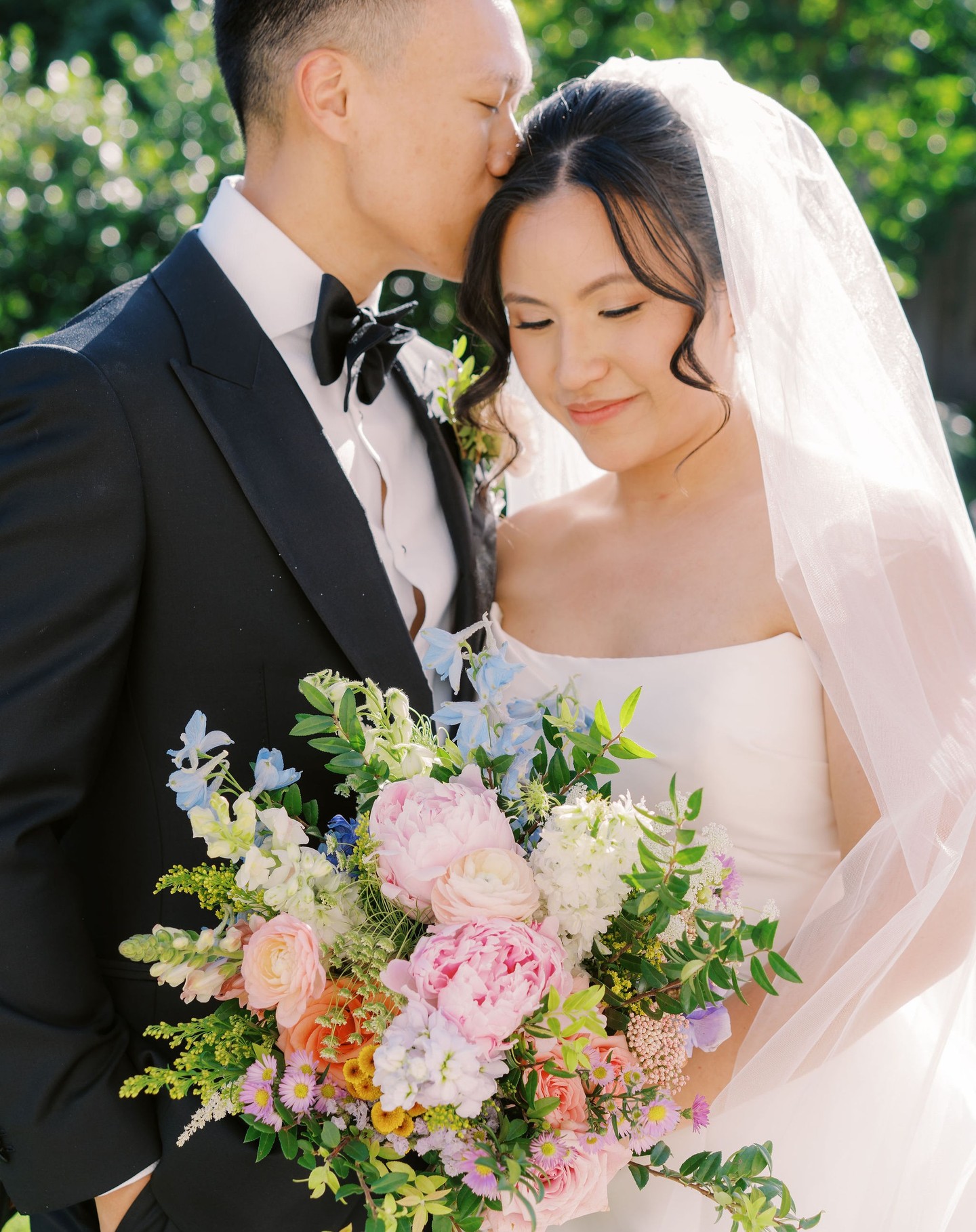 Bride holding lush bouquet of pink peonies, roses, blue delphiniums, and greenery with groom kissing her forehead