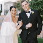 Bride in strapless ballgown and groom in black tuxedo with bow tie pose in front of colonial-style building