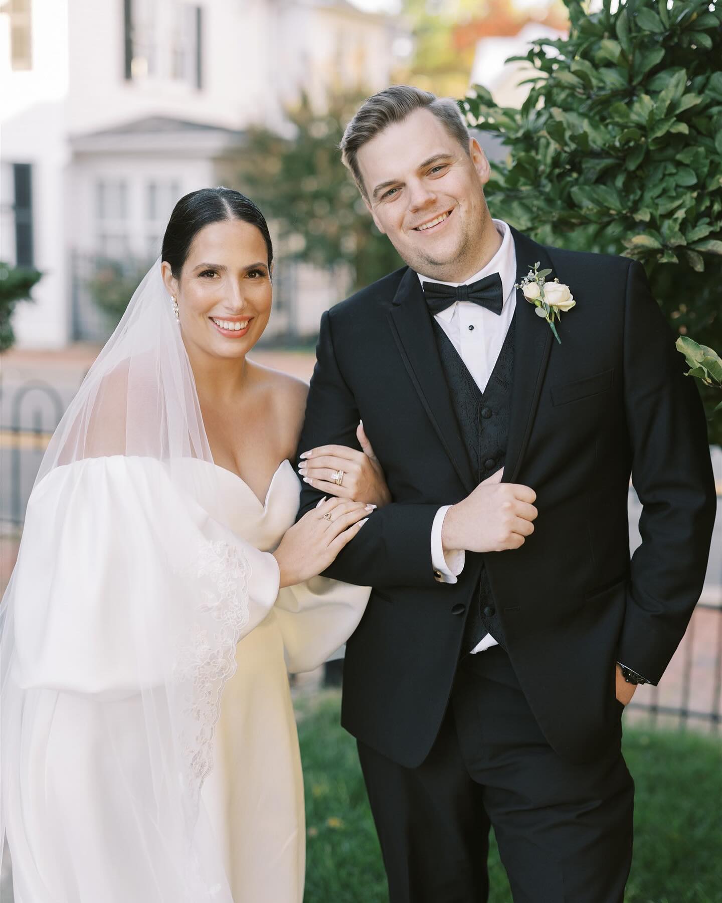 Bride in strapless ballgown and groom in black tuxedo with bow tie pose in front of colonial-style building
