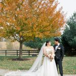 Bride and groom stand together under autumn tree with golden foliage and farm fence in background