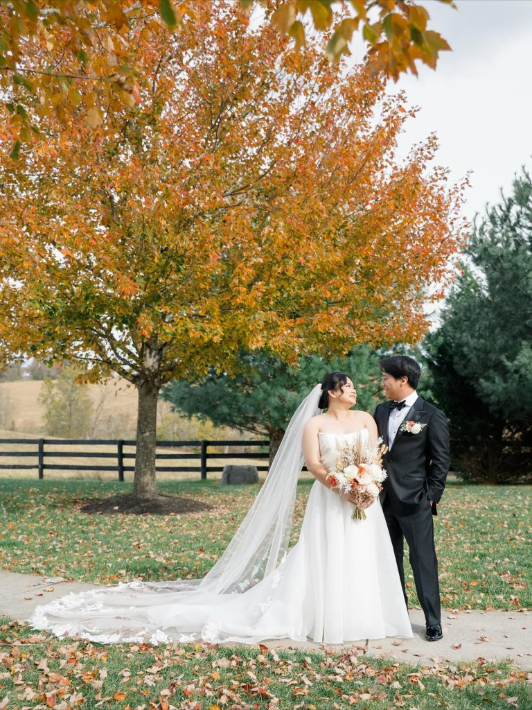 Bride and groom stand together under autumn tree with golden foliage and farm fence in background