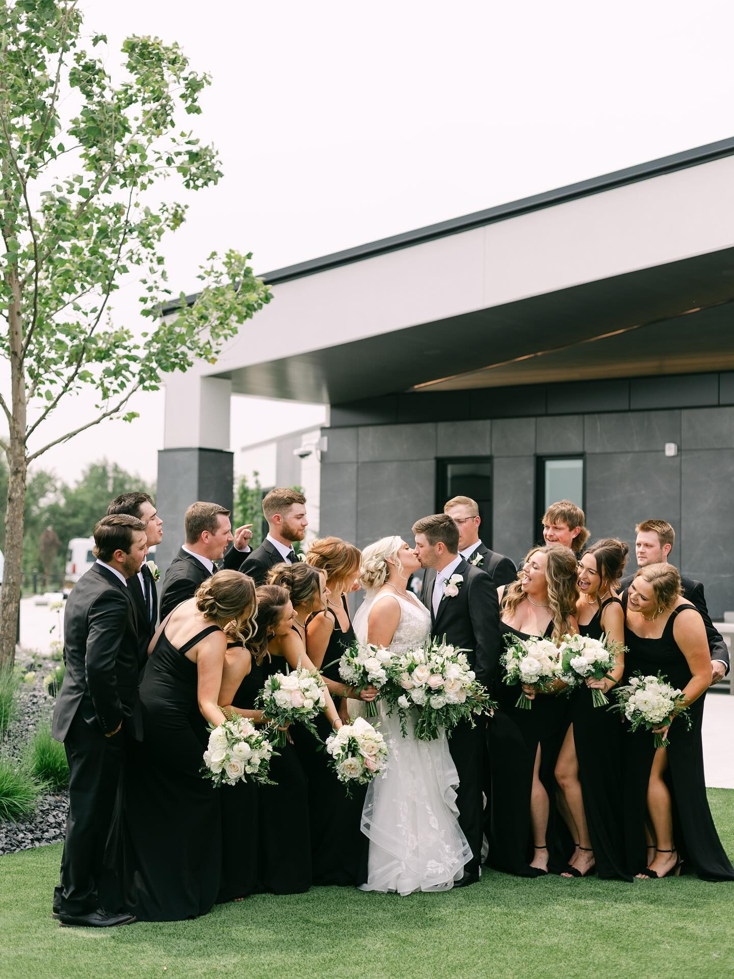 Wedding party gathered around bride and groom at contemporary venue with modern architecture and green lawn