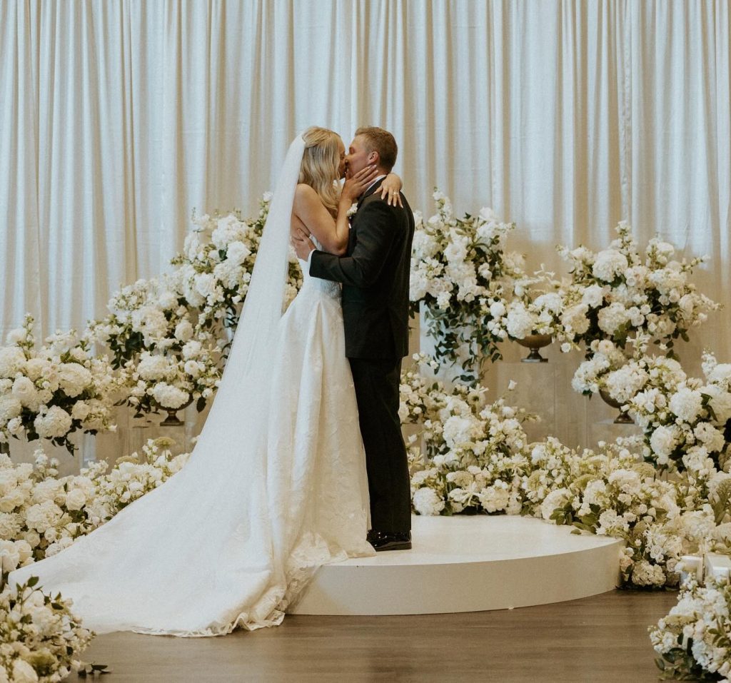 Newlyweds sharing their first kiss on elevated platform surrounded by lush white floral arrangements
