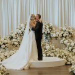 Newlyweds sharing their first kiss on elevated platform surrounded by lush white floral arrangements