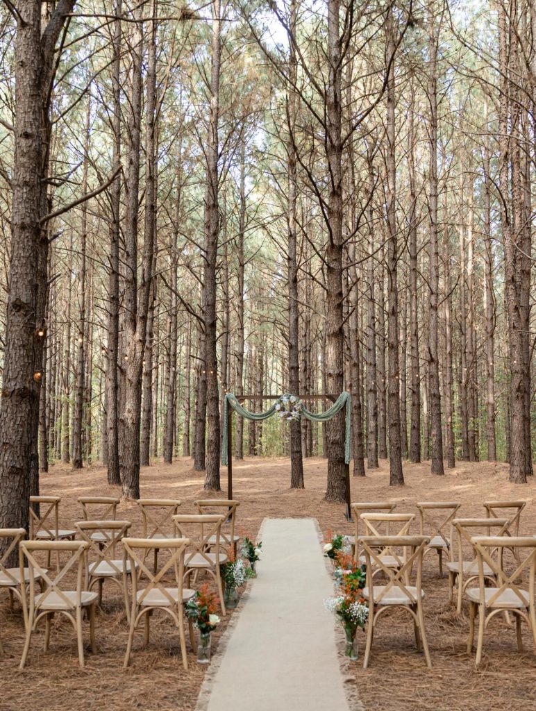 Woodland ceremony setup with wooden benches lining a white aisle runner among pine trees