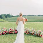 Bride in strapless lace wedding gown holding colorful bouquet, viewed from behind overlooking rolling green fields with vibrant flower installation