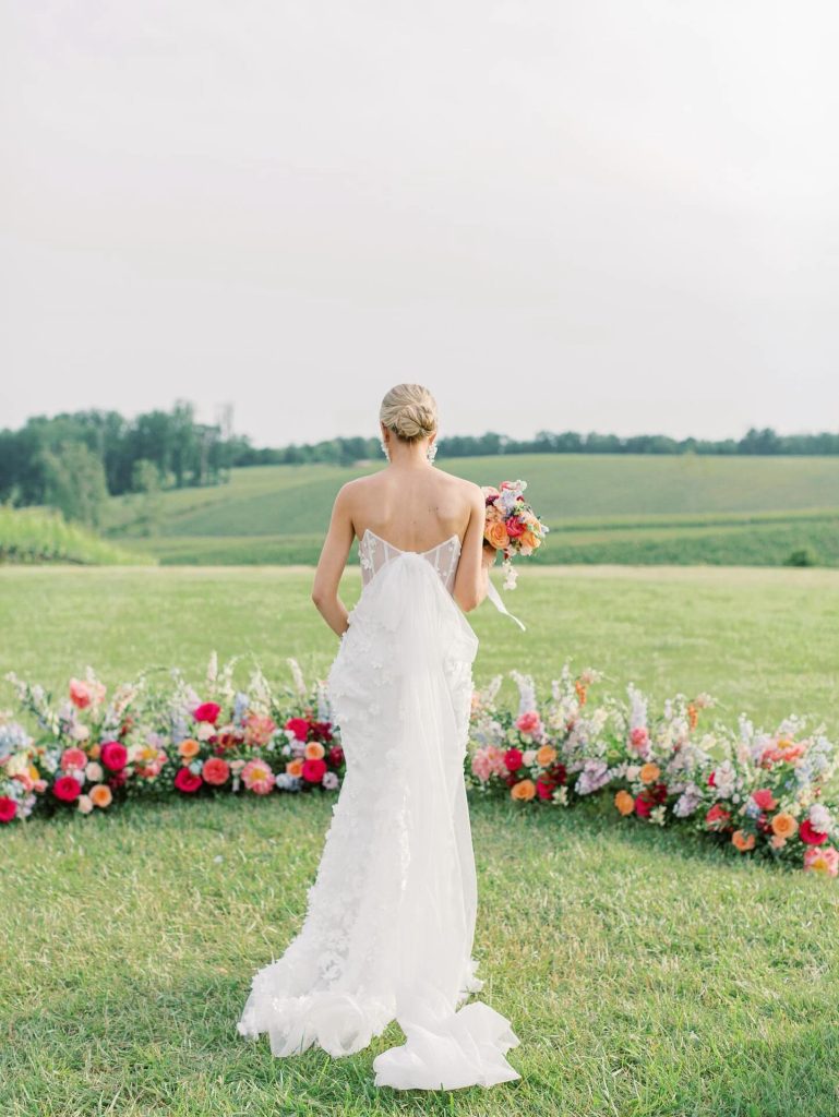 Bride in strapless lace wedding gown holding colorful bouquet, viewed from behind overlooking rolling green fields with vibrant flower installation