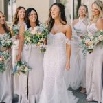Bride in strapless lace gown leads bridesmaids in champagne dresses holding pastel bouquets outdoors