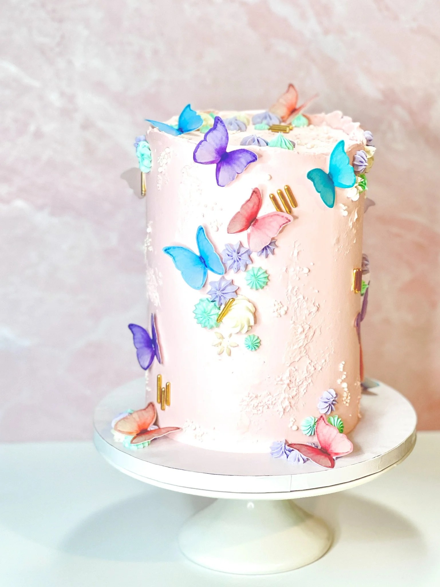 Single-tier wedding cake decorated with colorful edible butterflies, small flowers, and gold accents by Crumb Coat & Chill