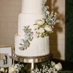 Three-tier white wedding cake with vertical line texture, adorned with eucalyptus and ranunculus flowers on gold stand