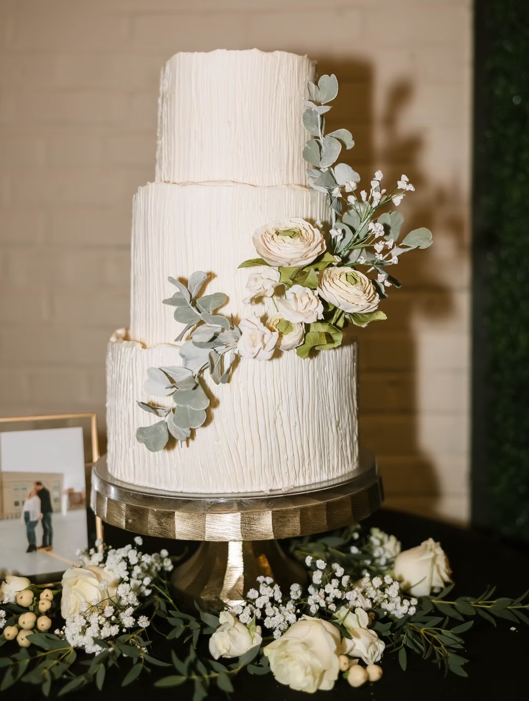 Three-tier white wedding cake with vertical line texture, adorned with eucalyptus and ranunculus flowers on gold stand