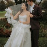Bride in elegant white gown with flowing veil and groom in burgundy suit pose in romantic outdoor garden setting