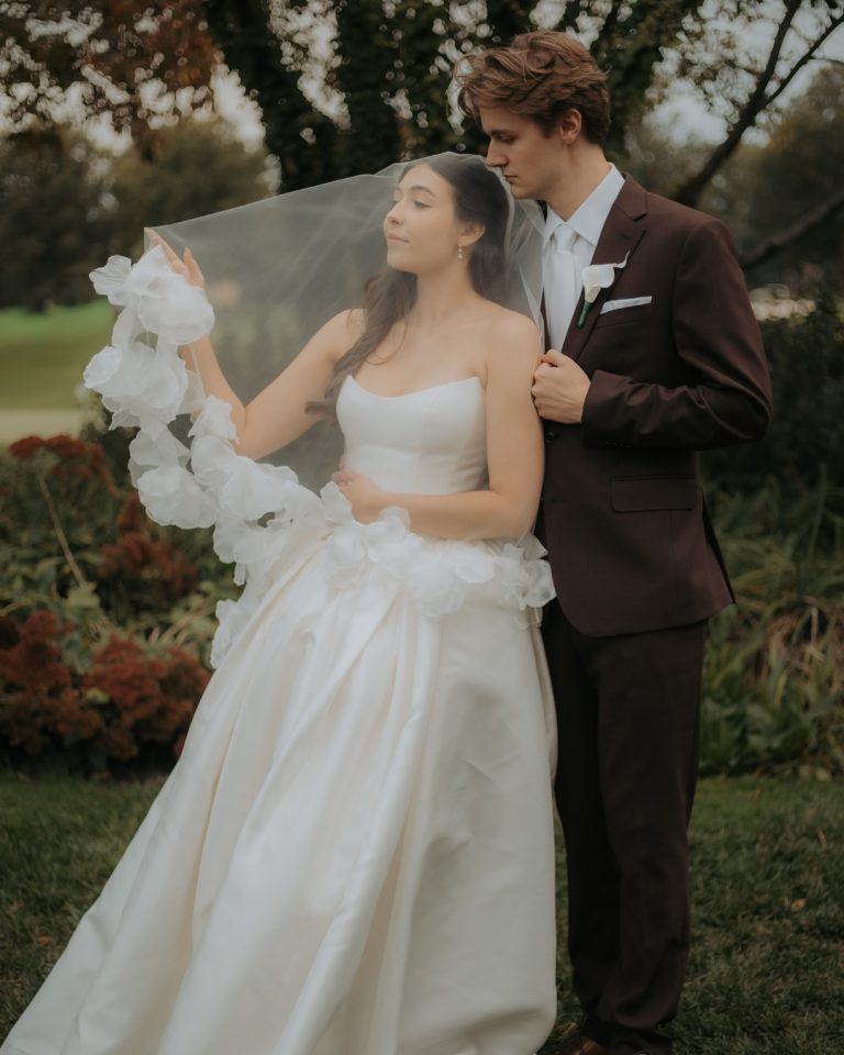 Bride in elegant white gown with flowing veil and groom in burgundy suit pose in romantic outdoor garden setting