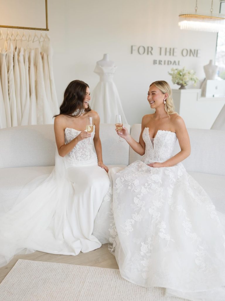 Two brides in elegant white ball gowns toasting with champagne in a luxury bridal salon