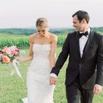 Bride in strapless lace gown and groom in black tuxedo walking through vineyard holding hands