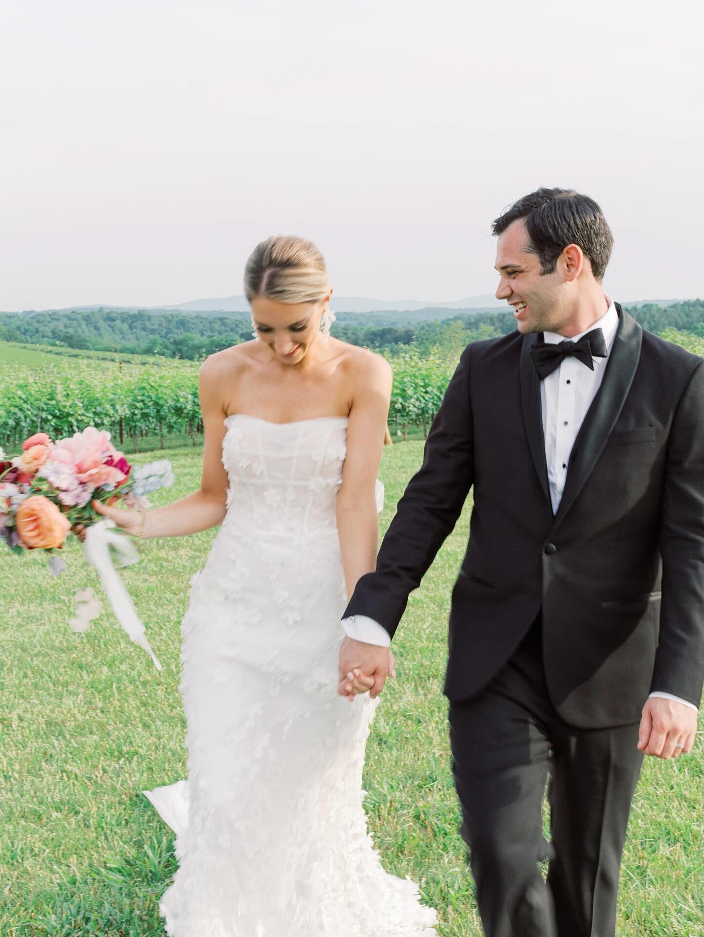 Bride in strapless lace gown and groom in black tuxedo walking through vineyard holding hands