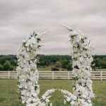 Curved white floral ceremony arch with flower-lined aisle on grassy lawn overlooking countryside