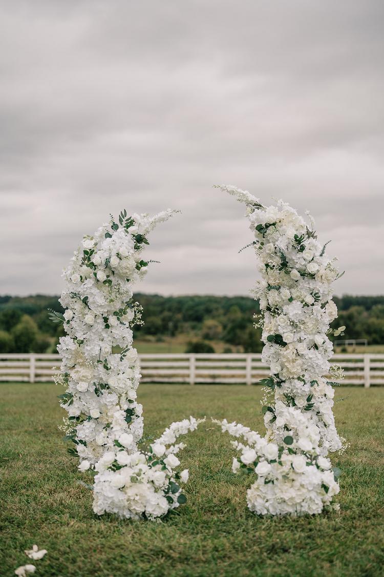 Curved white floral ceremony arch with flower-lined aisle on grassy lawn overlooking countryside