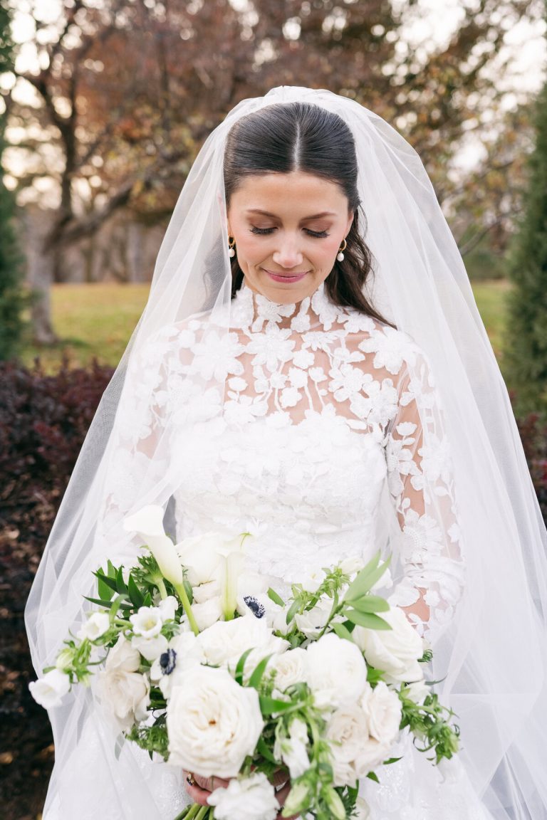 Bride in lace wedding dress and veil holding white bouquet with roses, calla lilies, and anemones