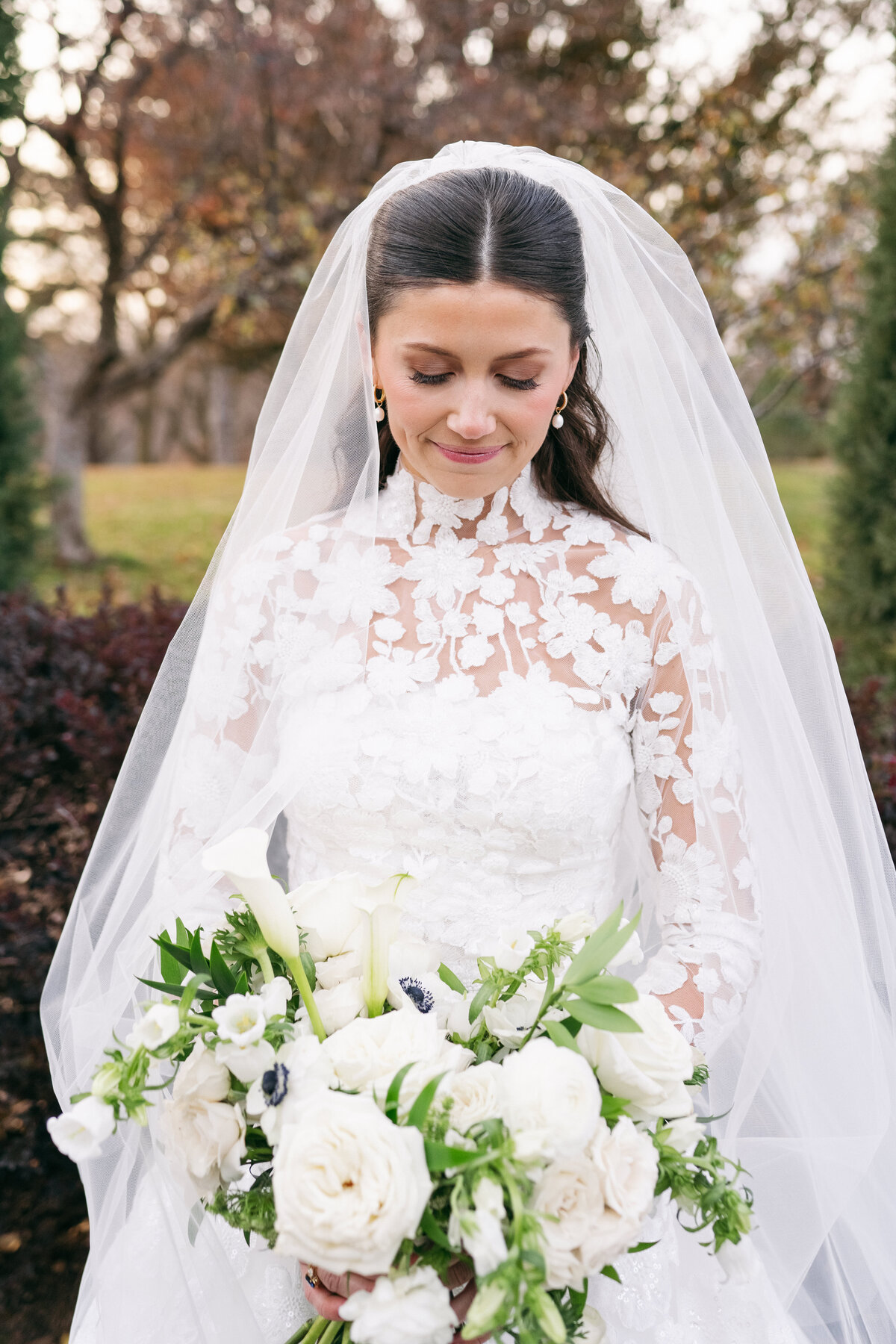 Bride in lace wedding dress and veil holding white bouquet with roses, calla lilies, and anemones