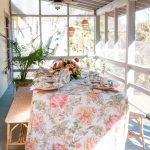 Screened porch dining table dressed with bold botanical tablecloth, coral floral centerpiece, and rattan chairs overlooking garden