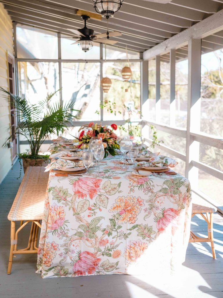 Screened porch dining table dressed with bold botanical tablecloth, coral floral centerpiece, and rattan chairs overlooking garden