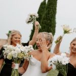 Bride in strapless gown reacting joyfully with hand to face while bridesmaids cheer with bouquets