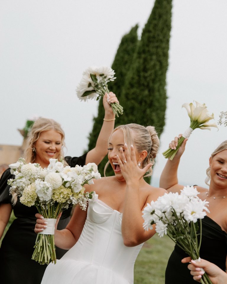 Bride in strapless gown reacting joyfully with hand to face while bridesmaids cheer with bouquets