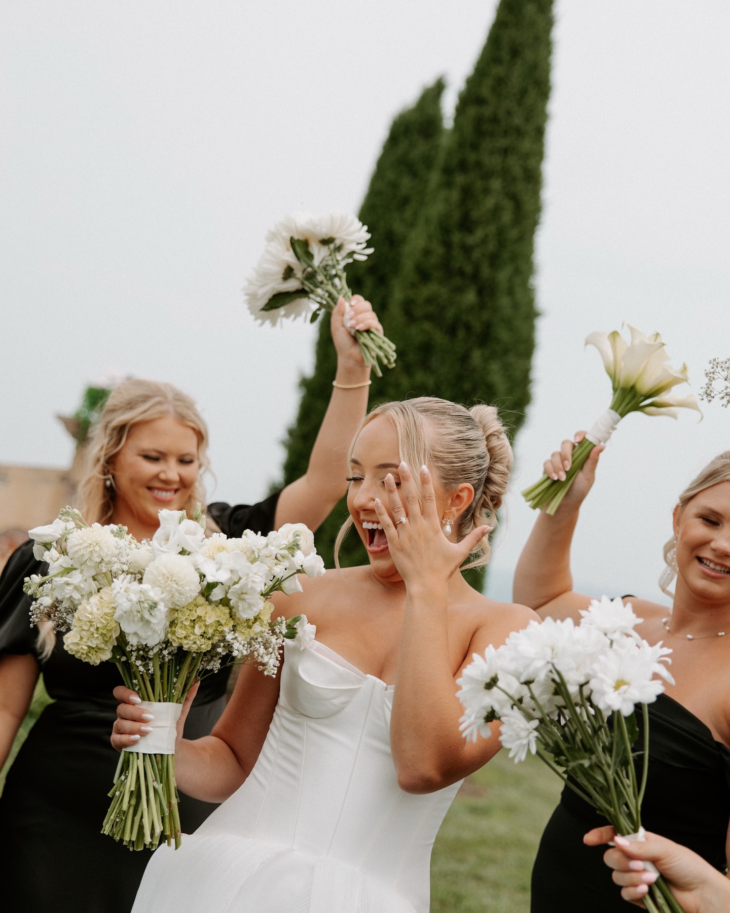 Bride in strapless gown reacting joyfully with hand to face while bridesmaids cheer with bouquets
