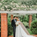 Bride in off-shoulder gown with cathedral train and groom in black tuxedo standing under flower-covered pergola