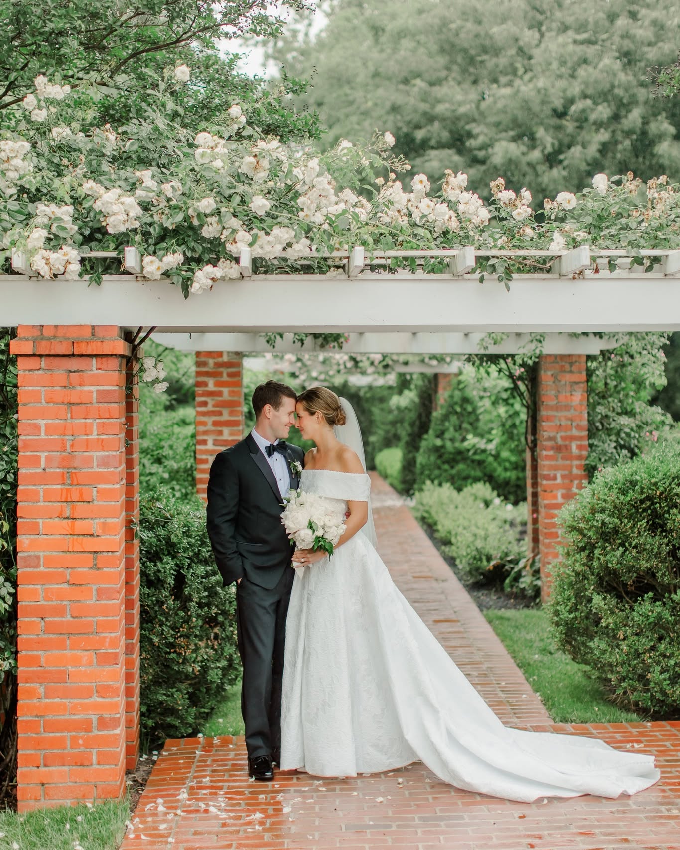Bride in off-shoulder gown with cathedral train and groom in black tuxedo standing under flower-covered pergola
