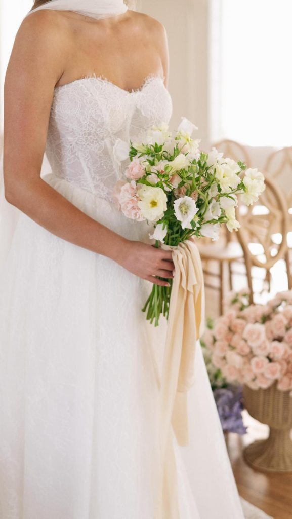 Bride in strapless lace wedding dress holding bouquet of white and pink flowers with silk ribbons