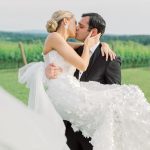 Groom carrying bride in floral-embellished wedding gown while kissing at vineyard with green hills beyond