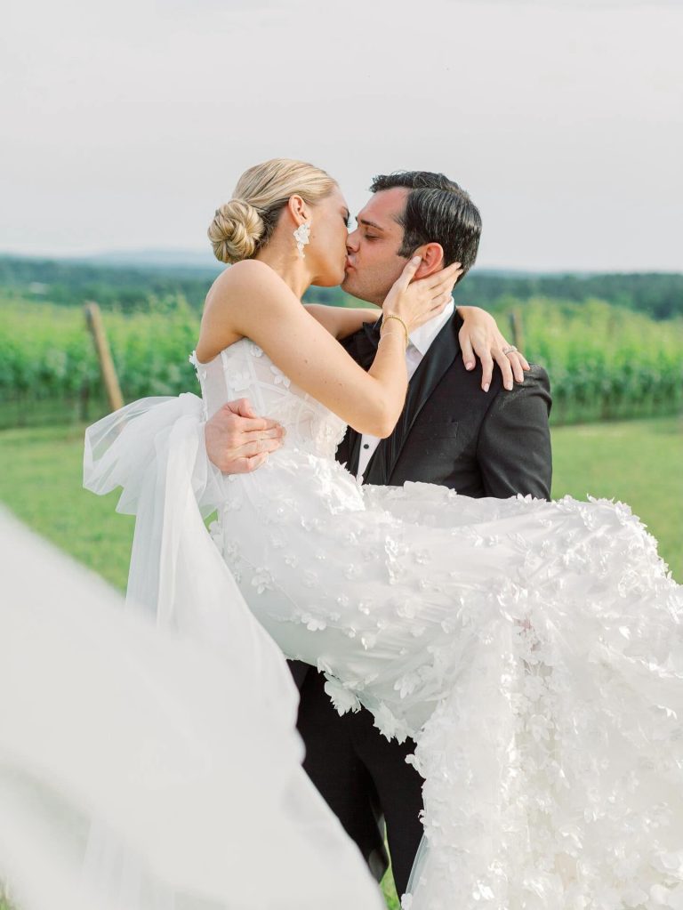 Groom carrying bride in floral-embellished wedding gown while kissing at vineyard with green hills beyond